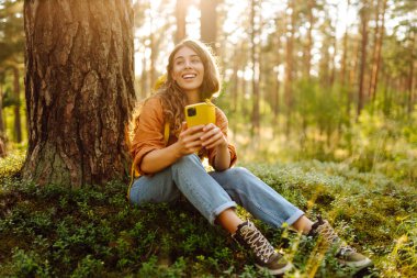 Genç bir kadın, sakin bir öğleden sonra güneşli bir ormanda akıllı telefonunu kullanırken bir ağaca yaslanır. Aktif yaşam tarzı, seyahat kavramı