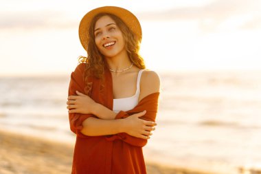 Genç bir kadın gün batımında kumsalda duruyor, güneşin sıcacık parıltısında güneşlenirken nazikçe saçlarına dokunuyor. Yaşam tarzı, turizm, doğa, aktif yaşam.