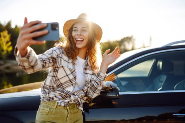 Parlak, güneşli bir ortamda park halindeki arabasının yanında akıllı telefonuyla selfie çekerken şapka takan genç bir kadın gülümsüyor ve el sallıyor. Etkin yaşam tarzı, blog yazma, doğa, hafta