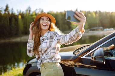 Parlak, güneşli bir ortamda park halindeki arabasının yanında akıllı telefonuyla selfie çekerken şapka takan genç bir kadın gülümsüyor ve el sallıyor. Etkin yaşam tarzı, blog yazma, doğa, hafta