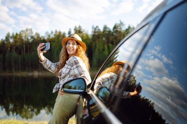 Parlak, güneşli bir ortamda park halindeki arabasının yanında akıllı telefonuyla selfie çekerken şapka takan genç bir kadın gülümsüyor ve el sallıyor. Etkin yaşam tarzı, blog yazma, doğa, hafta