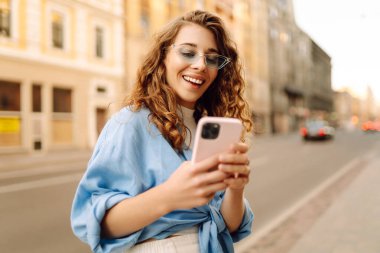 Şık güneş gözlüklü neşeli genç kadın akıllı telefonuyla meşgul oluyor, yeşillik dolu canlı bir şehir caddesinde güzel havanın tadını çıkarıyor. Teknoloji, bağlantı, blog yazma, modern yaşam