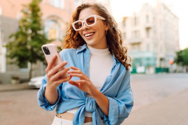 Şık güneş gözlüklü neşeli genç kadın akıllı telefonuyla meşgul oluyor, yeşillik dolu canlı bir şehir caddesinde güzel havanın tadını çıkarıyor. Teknoloji, bağlantı, blog yazma, modern yaşam