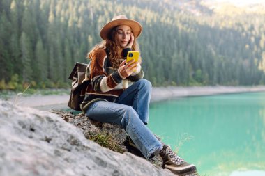 Samimi bir kazak ve şapka giyen genç bir kadın güzel bir gölün kenarındaki bir kayanın üzerinde oturuyor, gülümsüyor ve telefonunu kullanıyor. Macera, doğa, blog yazma.