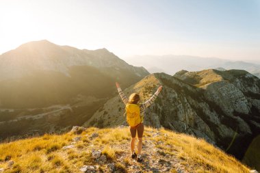 Neşeli genç bir kadın çimenli bir yamaçta oturur, güneş dağların ardında batarken gülümser. Yanında bir sırt çantası var ve bu güzel manzaranın tadını çıkarıyor. Yaşam tarzı, turizm.