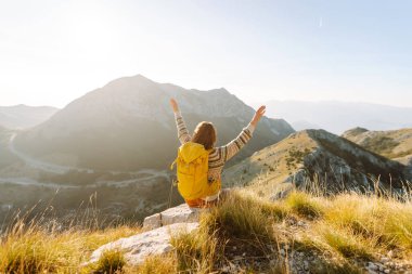 Neşeli genç bir kadın çimenli bir yamaçta oturur, güneş dağların ardında batarken gülümser. Yanında bir sırt çantası var ve bu güzel manzaranın tadını çıkarıyor. Yaşam tarzı, turizm.