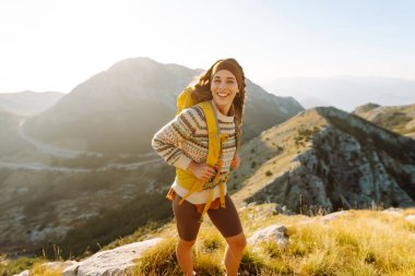 Neşeli genç bir kadın çimenli bir yamaçta oturur, güneş dağların ardında batarken gülümser. Yanında bir sırt çantası var ve bu güzel manzaranın tadını çıkarıyor. Yaşam tarzı, turizm.