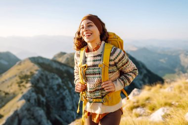 Neşeli genç bir kadın çimenli bir yamaçta oturur, güneş dağların ardında batarken gülümser. Yanında bir sırt çantası var ve bu güzel manzaranın tadını çıkarıyor. Yaşam tarzı, turizm.
