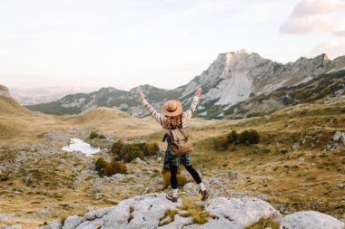 Geniş kenarlı şapkalı genç bir kadın, arkasında pitoresk bir manzara olan kayaların üzerinde otururken dağlarda güneşli bir öğleden sonranın tadını çıkarıyor. Çevre turizmi. Etkin yaşam biçimi.