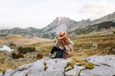 Geniş kenarlı şapkalı genç bir kadın, arkasında pitoresk bir manzara olan kayaların üzerinde otururken dağlarda güneşli bir öğleden sonranın tadını çıkarıyor. Çevre turizmi. Etkin yaşam biçimi.