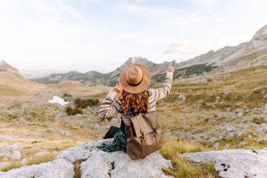 Geniş kenarlı şapkalı genç bir kadın, arkasında pitoresk bir manzara olan kayaların üzerinde otururken dağlarda güneşli bir öğleden sonranın tadını çıkarıyor. Çevre turizmi. Etkin yaşam biçimi.