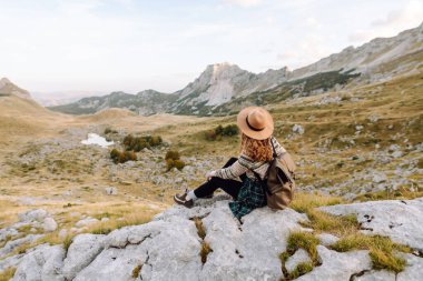 Geniş kenarlı şapkalı genç bir kadın, arkasında pitoresk bir manzara olan kayaların üzerinde otururken dağlarda güneşli bir öğleden sonranın tadını çıkarıyor. Çevre turizmi. Etkin yaşam biçimi.