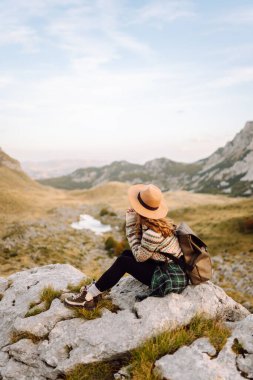 Geniş kenarlı şapkalı genç bir kadın, arkasında pitoresk bir manzara olan kayaların üzerinde otururken dağlarda güneşli bir öğleden sonranın tadını çıkarıyor. Çevre turizmi. Etkin yaşam biçimi.