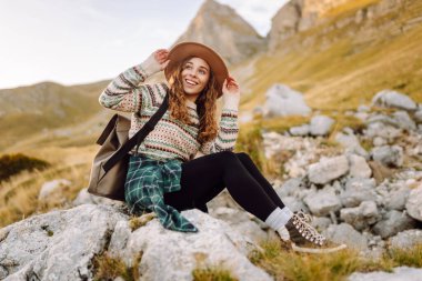 Geniş kenarlı şapkalı genç bir kadın, arkasında pitoresk bir manzara olan kayaların üzerinde otururken dağlarda güneşli bir öğleden sonranın tadını çıkarıyor. Çevre turizmi. Etkin yaşam biçimi.