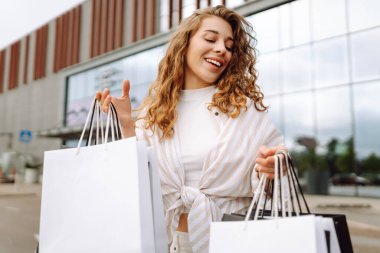 Genç bir kadın güneşli bir günde modern bir perakende mağazasının önünde alışveriş torbalarını neşeyle tutuyor. Satış, alışverişler, alışveriş, yaşam tarzı konsepti.