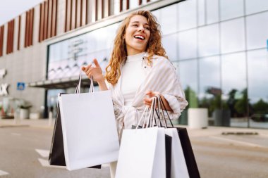 Genç bir kadın güneşli bir günde modern bir perakende mağazasının önünde alışveriş torbalarını neşeyle tutuyor. Satış, alışverişler, alışveriş, yaşam tarzı konsepti.