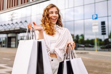 Genç bir kadın güneşli bir günde modern bir perakende mağazasının önünde alışveriş torbalarını neşeyle tutuyor. Satış, alışverişler, alışveriş, yaşam tarzı konsepti.
