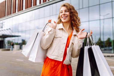 Genç bir kadın güneşli bir günde modern bir perakende mağazasının önünde alışveriş torbalarını neşeyle tutuyor. Satış, alışverişler, alışveriş, yaşam tarzı konsepti.