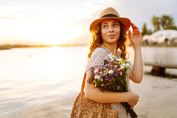 Молодая женщина, держа букет полевых цветов на берегу реки во время заката, запечатлевает красоту природы и радость момента