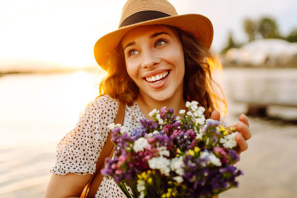 Молодая женщина, держа букет полевых цветов на берегу реки во время заката, запечатлевает красоту природы и радость момента