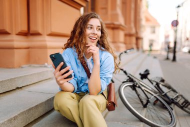 Genç bir kadın, güneşli bir öğleden sonra yürüyüşü sırasında şehirde bisiklete yaslanırken telefonunu kontrol ediyor. Selfie zamanı. İnsanlar, boş zaman, blog ve yaşam tarzı