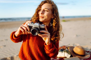 Genç bir kadın, öğleden sonra saatlerinde sıcak bir ışıkla fotoğraf çekerken kumsalda güneşli bir günün tadını çıkarıyor. Seyahat, blog, teknoloji kavramı.