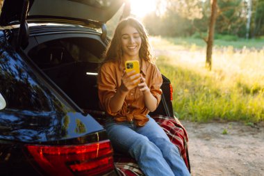 Genç bir kadın açık havada güneşli bir günün tadını çıkarırken arabasında oturuyor ve gün batımında akıllı telefonuyla iletişim kuruyor. Seyahat, hafta sonu, doğa, rahatlama ve yaşam tarzı konsepti