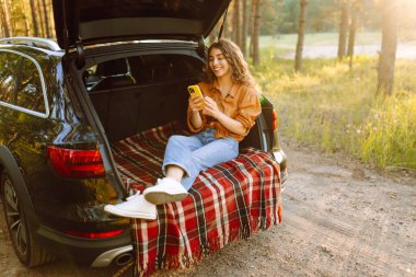 Genç bir kadın açık havada güneşli bir günün tadını çıkarırken arabasında oturuyor ve gün batımında akıllı telefonuyla iletişim kuruyor. Seyahat, hafta sonu, doğa, rahatlama ve yaşam tarzı konsepti