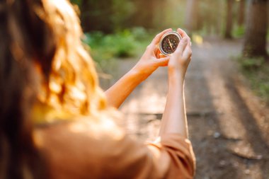 Kıvırcık saçlı bir yürüyüşçü, elinde bir pusula tutarak uzun ağaçlar, yeşillikler ve sıcak öğleden sonra ışığıyla dolu güneşli bir ormanda dikkatlice gezinir..