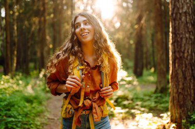 Kıvırcık saçlı, uzun ağaçlar ve sıcak güneş ışığıyla çevrili sarı bir sırt çantası ile orman yolunda yürüyen mutlu bir genç kadın. Seyahatler. Aktif yaşam tarzı. Yürüyüş.