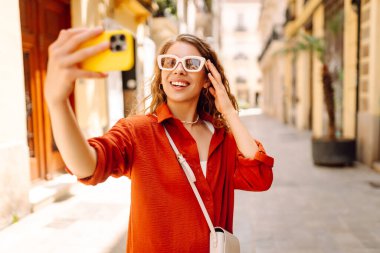 Gündüz vakti güneşli bir caddede yürürken şık güneş gözlükleriyle selfie çeken neşeli bir kadın. Seyahat, turizm, teknoloji, blog, iletişim kavramı.