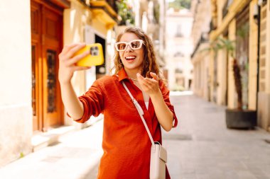 Gündüz vakti güneşli bir caddede yürürken şık güneş gözlükleriyle selfie çeken neşeli bir kadın. Seyahat, turizm, teknoloji, blog, iletişim kavramı.