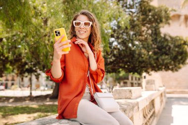 Gündüz vakti güneşli bir caddede yürürken şık güneş gözlükleriyle selfie çeken neşeli bir kadın. Seyahat, turizm, teknoloji, blog, iletişim kavramı.