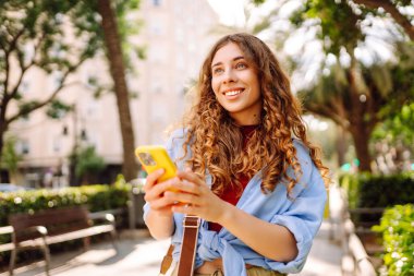 Gündüz vakti palmiye ağaçlarıyla kaplı güneşli bir sokakta yürürken genç bir kadın telefon kullanıyor. İş, teknoloji, blog, iletişim kavramı.