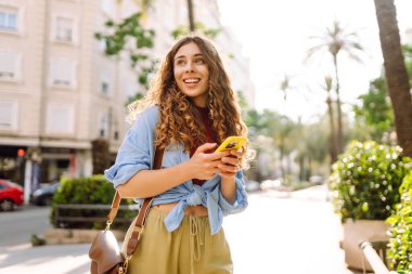 Gündüz vakti palmiye ağaçlarıyla kaplı güneşli bir sokakta yürürken genç bir kadın telefon kullanıyor. İş, teknoloji, blog, iletişim kavramı.