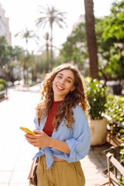 Gündüz vakti palmiye ağaçlarıyla kaplı güneşli bir sokakta yürürken genç bir kadın telefon kullanıyor. İş, teknoloji, blog, iletişim kavramı.
