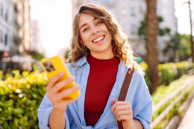 Gündüz vakti palmiye ağaçlarıyla kaplı güneşli bir sokakta yürürken genç bir kadın telefon kullanıyor. İş, teknoloji, blog, iletişim kavramı.