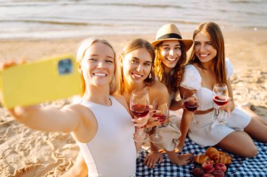 Okyanus kenarında güneşli bir öğleden sonra selfie çekerken ve içki içerken sahilde toplanan dört güzel bayan arkadaş. - Evet. Tatiller, doğa, rahatlama konsepti.