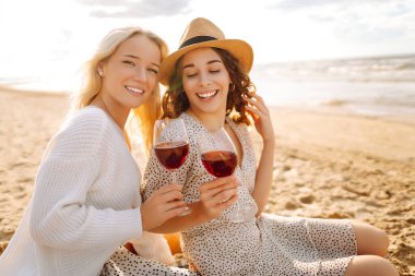 Gün batımında kumlu bir plajda kırmızı şarap içen iki kadın arkadaş kahkaha ve sıcaklıkla dolu neşeli bir anı yakalıyorlar..