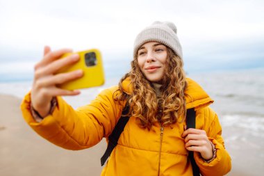 Neşeli genç bir kadın bulutlu bir günde kumlu bir plajda selfie çekiyor ve arka planda dalgalar çarpışıyor. Selfie zamanı. Soğuk rüzgarlı hava. Açık havada. Seyahat kavramı.