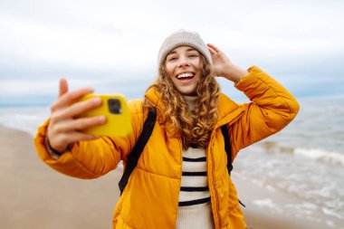 Neşeli genç bir kadın bulutlu bir günde kumlu bir plajda selfie çekiyor ve arka planda dalgalar çarpışıyor. Selfie zamanı. Soğuk rüzgarlı hava. Açık havada. Seyahat kavramı.