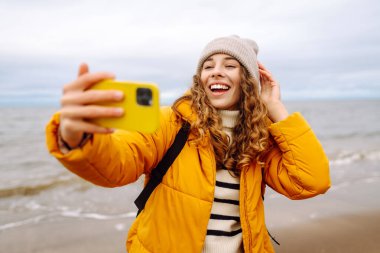 Neşeli genç bir kadın bulutlu bir günde kumlu bir plajda selfie çekiyor ve arka planda dalgalar çarpışıyor. Selfie zamanı. Soğuk rüzgarlı hava. Açık havada. Seyahat kavramı.