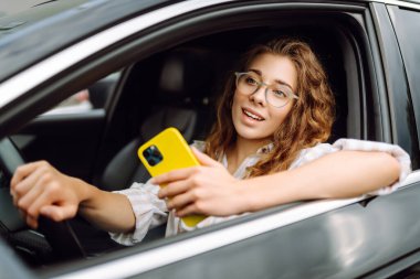Kıvırcık saçlı genç bir kadın güneşli bir günde şehir caddesinde araba kullanırken telefon kullanıyor. Boş zaman, seyahat, teknoloji. Araba paylaşımı.