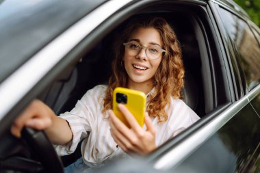 Kıvırcık saçlı genç bir kadın güneşli bir günde şehir caddesinde araba kullanırken telefon kullanıyor. Boş zaman, seyahat, teknoloji. Araba paylaşımı.