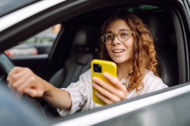 Kıvırcık saçlı genç bir kadın güneşli bir günde şehir caddesinde araba kullanırken telefon kullanıyor. Boş zaman, seyahat, teknoloji. Araba paylaşımı.