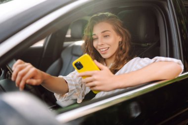 Kıvırcık saçlı genç bir kadın güneşli bir günde şehir caddesinde araba kullanırken telefon kullanıyor. Boş zaman, seyahat, teknoloji. Araba paylaşımı.