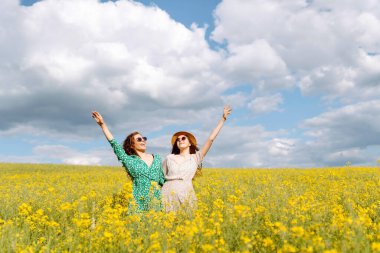 İki arkadaş canlı sarı bir çiçek tarlasında şık kıyafetler ve güneş gözlüğü takarak güneşli bir günün tadını çıkarıyorlar. Doğa, tatil, rahatlama ve yaşam tarzı. Yaz manzarası.