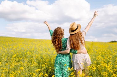 İki arkadaş canlı sarı bir çiçek tarlasında şık kıyafetler ve güneş gözlüğü takarak güneşli bir günün tadını çıkarıyorlar. Doğa, tatil, rahatlama ve yaşam tarzı. Yaz manzarası.