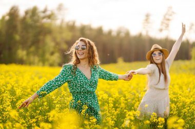 İki arkadaş canlı sarı bir çiçek tarlasında şık kıyafetler ve güneş gözlüğü takarak güneşli bir günün tadını çıkarıyorlar. Doğa, tatil, rahatlama ve yaşam tarzı. Yaz manzarası.