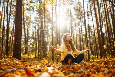 Bir kadın, sonbaharda yapraklar ve uzun ağaçlarla çevrili güneşli bir ormanda yoga yapıyor. Heather yaşam tarzı.